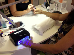 Esteticista realizando la técnica de esmalte permanente (EP) en una mesa de trabajo con dos lámparas, una a cada lado de la clienta. Obsérvese como el primer dedo de la mano introducida en la lámpara se encuentra fuera de ella. Para que el acrilato polimerice correctamente se debe exponer este dedo a la luz independientemente, en un segundo tiempo.