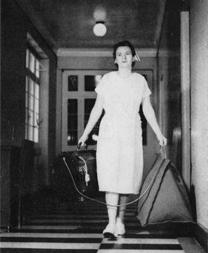 Enfermera transportando un recién nacido prematuro. En su mano derecha lleva al neonato en una caja de madera con ventana y en la mano izquierda una bolsa con el oxígeno. Hospital Calvo Mackenna, década de los años 60.