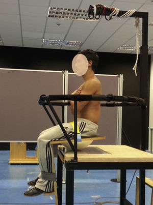 Vista lateral de un participante durante la ejecución de una prueba sobre asiento inestable. El asiento está apoyado sobre una plataforma de fuerzas que permite el análisis del desplazamiento del centro de presiones y de esta forma, la valoración del control postural del tronco en sedestación. Una barandilla de seguridad rodea al participante para evitar caídas y hacer que este se sienta seguro durante la prueba.